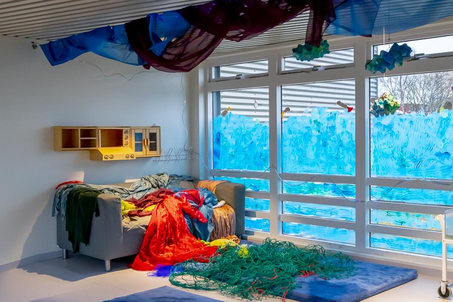 A corner at Lyngheimar preschool decorated as an ocean floor. Sofa, rug, fishing net, streamers on the ceiling and blue-painted window.