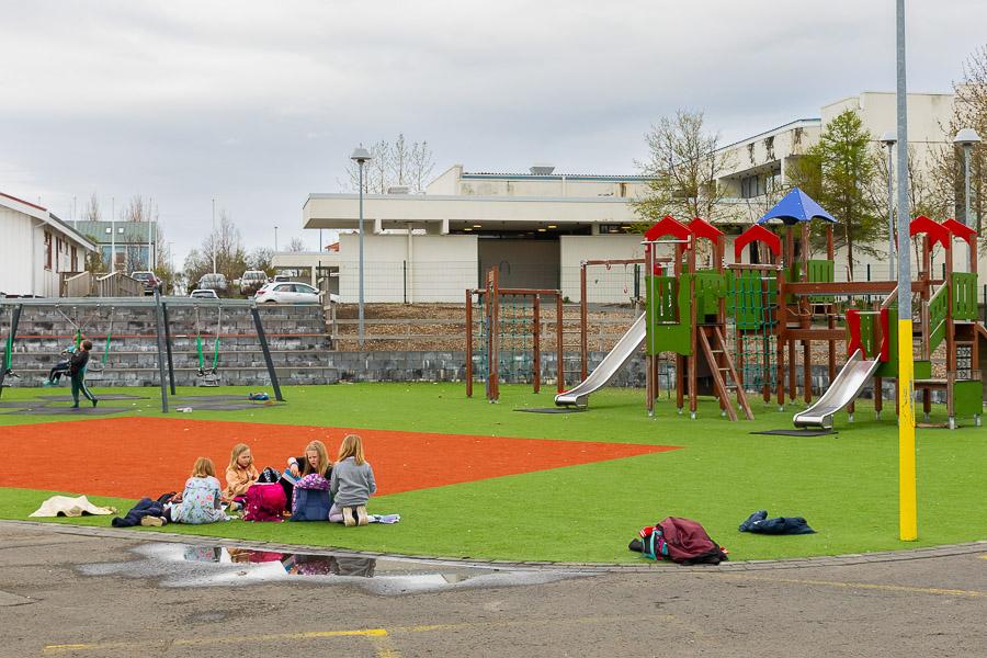An outdoor play area with a jungle gym, swings, and a playground castle. Children sit in a group on a green surface. 