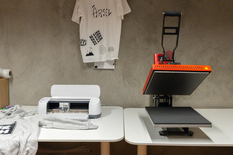 White tables with a t-shirt printing station. A t-shirt with "Ársel" lettering hangs on a wall. 
