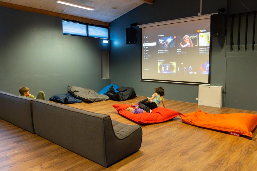 A hall with a large screen and gray, multicolored cushions. Children watching a screen.