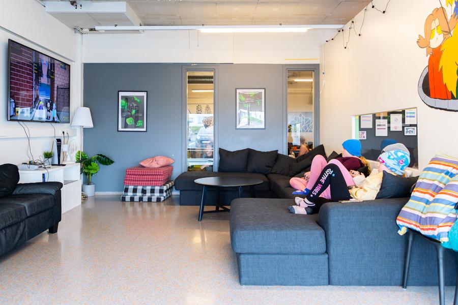 Children sit on a couch watching a wall screen. A small table sits by the couch with a stack of cushions. Pictures and wall decorations hang on the walls. 