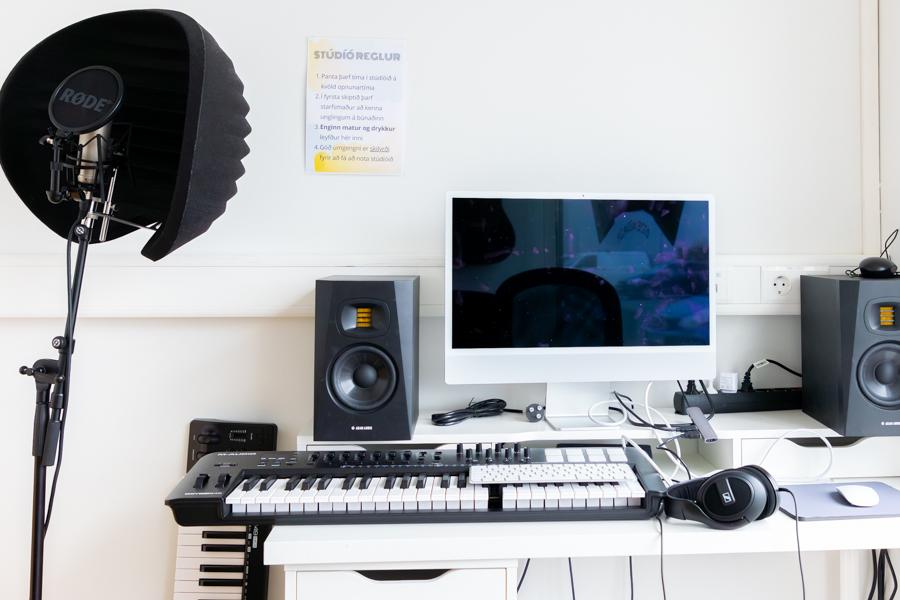 Table with computer monitor, keyboard, speakers and other equipment. A sheet with studio rules is posted on the wall. 