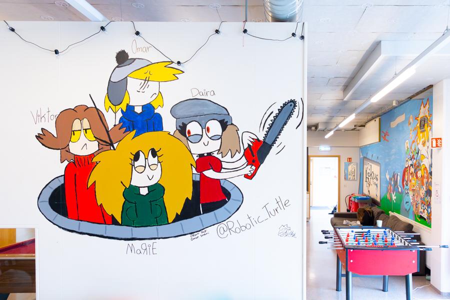 Hand-drawn wall artwork in the foreground. To the side, you can see a large decorated wall, couch and foosball table. 