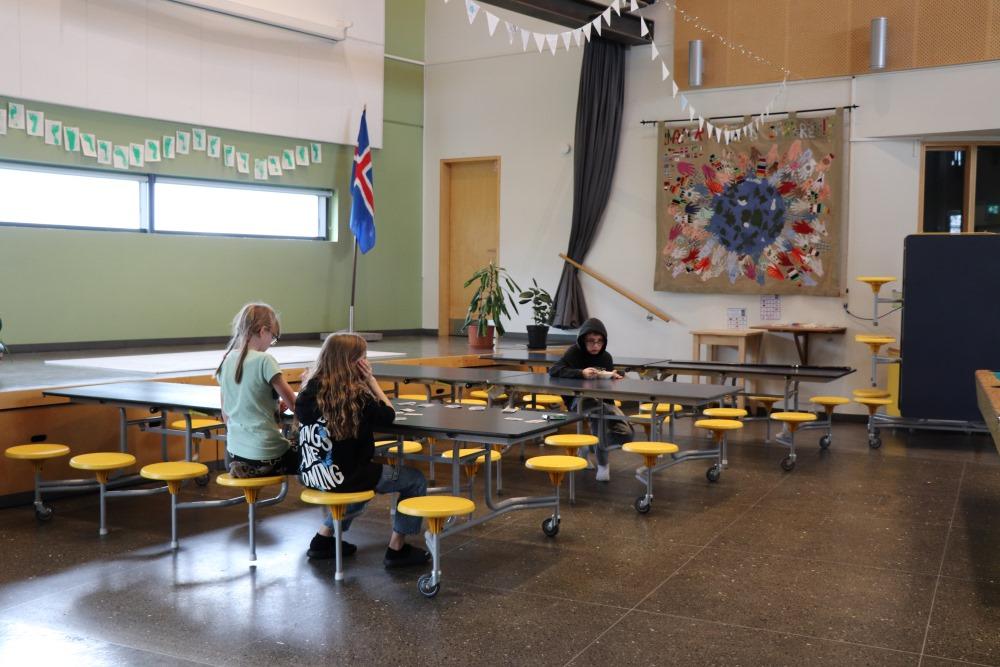 Langborð í röðum með gulum kollum. Börn að spila og borða við borðin. Fánar í lofti, íslenski fáninn til hliðar og mynd á vegg. 