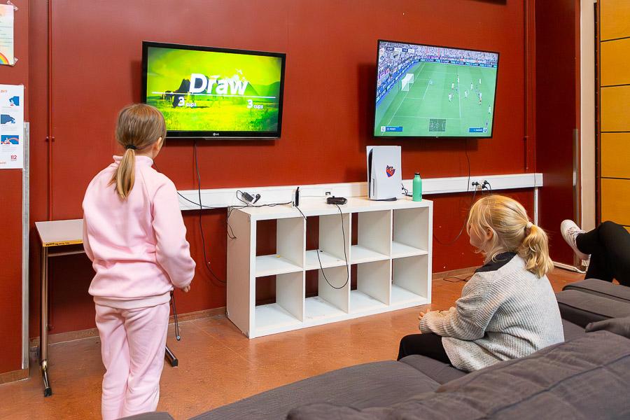 Children sitting and standing in front of screens against a red wall. 
