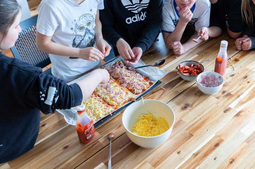 Börn standa við borð og eru að setja álegg á pizzu.