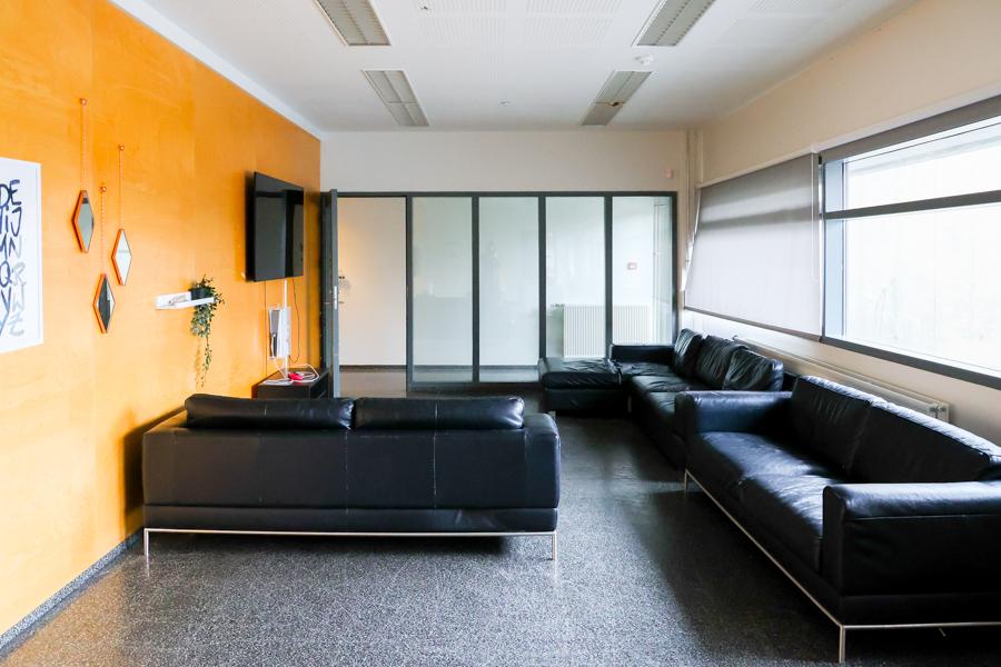 Room with windows on one side and an orange wall on the other. Screen on the wall with black couches arranged along the wall and by the screen. 