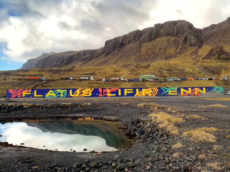 The mural Flatus still lives, at the base of Esja. A puddle in the foreground, Mount Esja in the background.