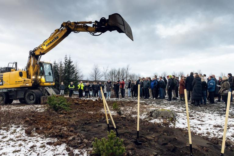 Hópur fólks hægra megin, í forgrunni fimm skóflur, vinstra megin stór grafa með skófluna á lofti yfir fólkinu. Frá fyrstu skóflustungu að verknámsaðstöðu í FB.