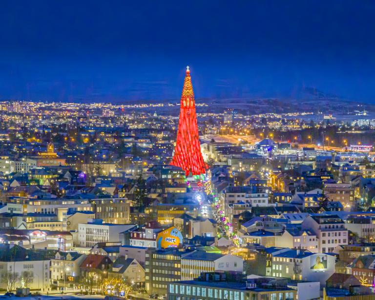 Aerial view of the city during the Winter Lights Festival. Evening view over Þingholt, with Hallgrímskirkja's illuminated tower glowing red against the sky.