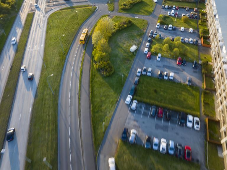 Loftmynd af Strætó að keyra á götu, innan um grænan gróður. Bílastæði til hægri á mynd.