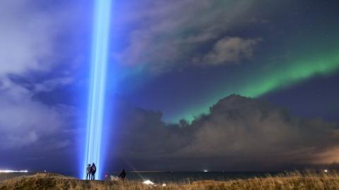 Fólk stendur við Friðarsúluna í Viðey með norðurljós á himni.