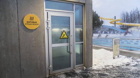 Sauna at Breiðholtslaug, showing the exterior, entrance door, and sign next to the door. Swimming pool in the background. Winter, snow.