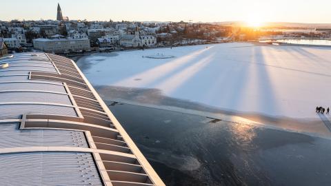 The roof of City Hall in the foreground, the frozen Tjörnin pond, the sun rising in the distance. Hallgrímskirkja church and Þingholt in the background.
