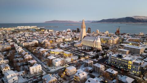 Aerial view over the city center, with Hallgrímskirkja prominent in the center, and Faxaflói Bay and the mountains in the distance. Clear skies, winter light.