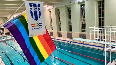 The Rainbow Flag and the Reykjavík flag hang inside the Sundhöllin swimming pool, with the diving board and pool visible in the background
